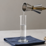 A metal fluid-ounce jigger pours water into an unnumbered graduated glass cylinder